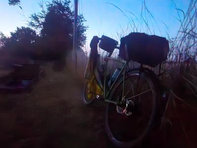 Touring bicycle with black front and rear carriers, yellow jerkin hanging nearby, parked outdoors at dawn in misty graveyard with a field of tall grass framing the right side.