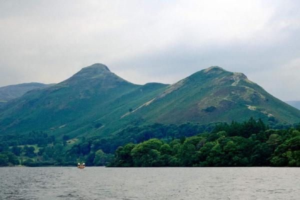 Cat Bells seen from across Derwent Water, with its twin summits rising above a band of dark green forest along the shoreline. A small motorboat sits on the rippling grey water in the foreground, and the hills are covered in a mix of grass and bracken under an overcast sky.