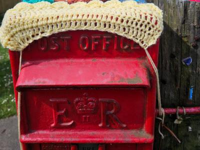 Red Royal Mail post box decorated with yarn bombing. A pale yellow crochet trim covers the 'Post Office' lettering on the box door. A large white crochet snail with tall stalk eyes and brown eyebrows perches on top. Residential street visible in background.