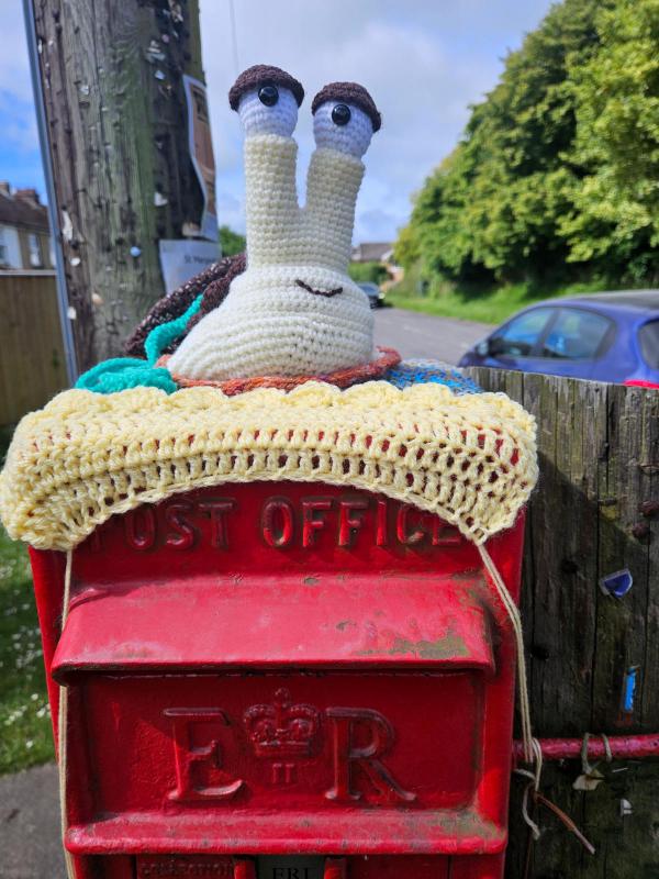 Red Royal Mail post box decorated with yarn bombing. A pale yellow crochet trim covers the &lsquo;Post Office&rsquo; lettering on the box door. A large white crochet snail with tall stalk eyes and brown eyebrows perches on top. Residential street visible in background.