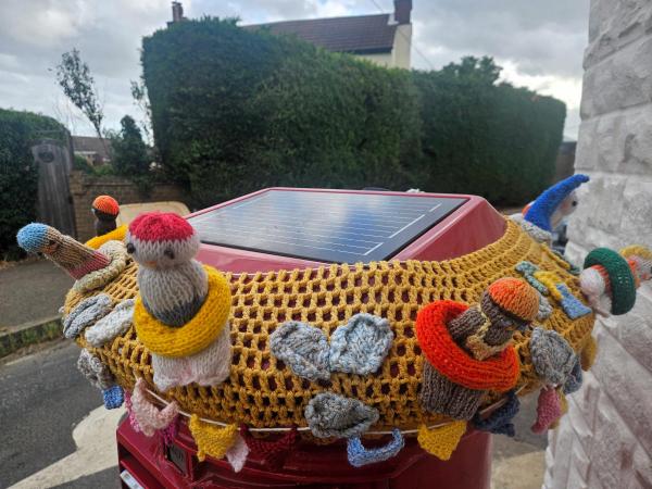 Red UK pillar box covered in mustard-yellow knitted mesh with handmade knitted figurines wearing swimming costumes and bright rubber rings. A solar panel sits on top. Residential street background.