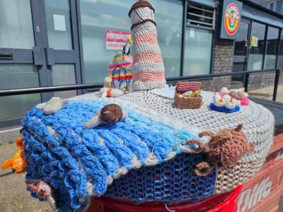 Crocheted seaside scene on red postbox featuring striped lighthouse, blue waves, octopus, seal, picnic basket and nautical decorations.