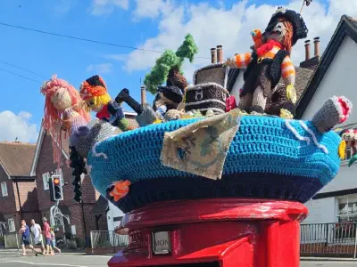 A pirate-themed knitted scene atop a red post box. The display features multiple pirates, a treasure chest, a shark, a palm tree, and a mermaid with pink hair, all crafted with yarn and arranged on a blue crocheted base, with houses and a traffic light in the background.
